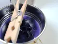 Proceso para como hacer velas de soja: cera morada enfriándose en un recipiente metálico, sujeta por pinzas de madera.