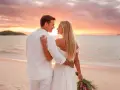 Pareja en la playa al atardecer, celebrando bodas románticas. Él, con camisa blanca, la abraza. Ella, con vestido de novia, mira al horizonte.