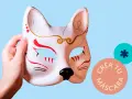 Mano sostiene un antifaz personalizado de zorro, con detalles rojos y dorados. Círculos con texto "Crea tu máscara" y un asterisco.