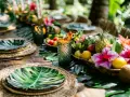 Mesa decorada para una fiesta hawaiana casera, con centros de mesa de frutas tropicales, flores y hojas de palma.