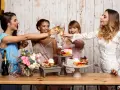 Cuatro amigas brindan con copas de champán en unas fiestas en casa, rodeadas de pasteles y flores.