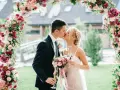 Novios besándose bajo un arco floral rosa y blanco, celebrando su boda barata con copas de champán.