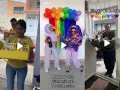 Una fiesta temática de colores vibrantes con globos arcoíris y atuendos morados. Un hombre entrega una canasta de regalos.