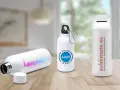 Tres bidones personalizados en una mesa de madera. Uno con logo "BAUHAUS", otro con "YOUR LOGO HERE" y el tercero con "sublimate.es".
