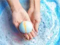 Manos sostienen una bomba de baño azul y amarilla sobre espuma burbujeante. ¡Perfecto para hacer bombas de baño caseras!