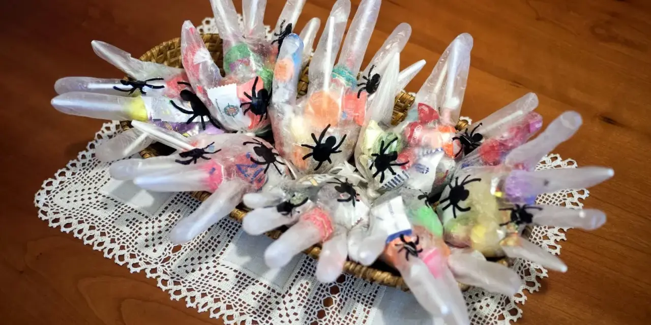 Manualidades chuches Halloween: guantes transparentes rellenos de caramelos y decorados con arañas negras, presentados en una cesta sobre un mantel de encaje.