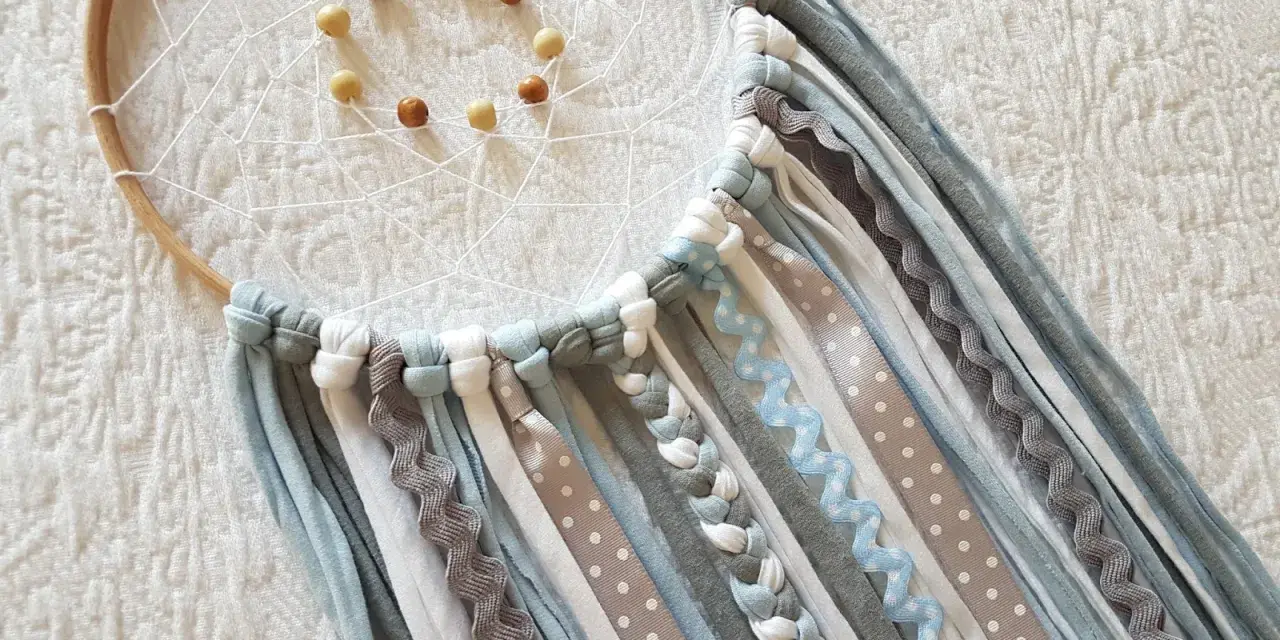 Un atrapasueños casero con aros de madera, red blanca y cuentas. Cintas de tela y encaje cuelgan en tonos grises, blancos y azules.