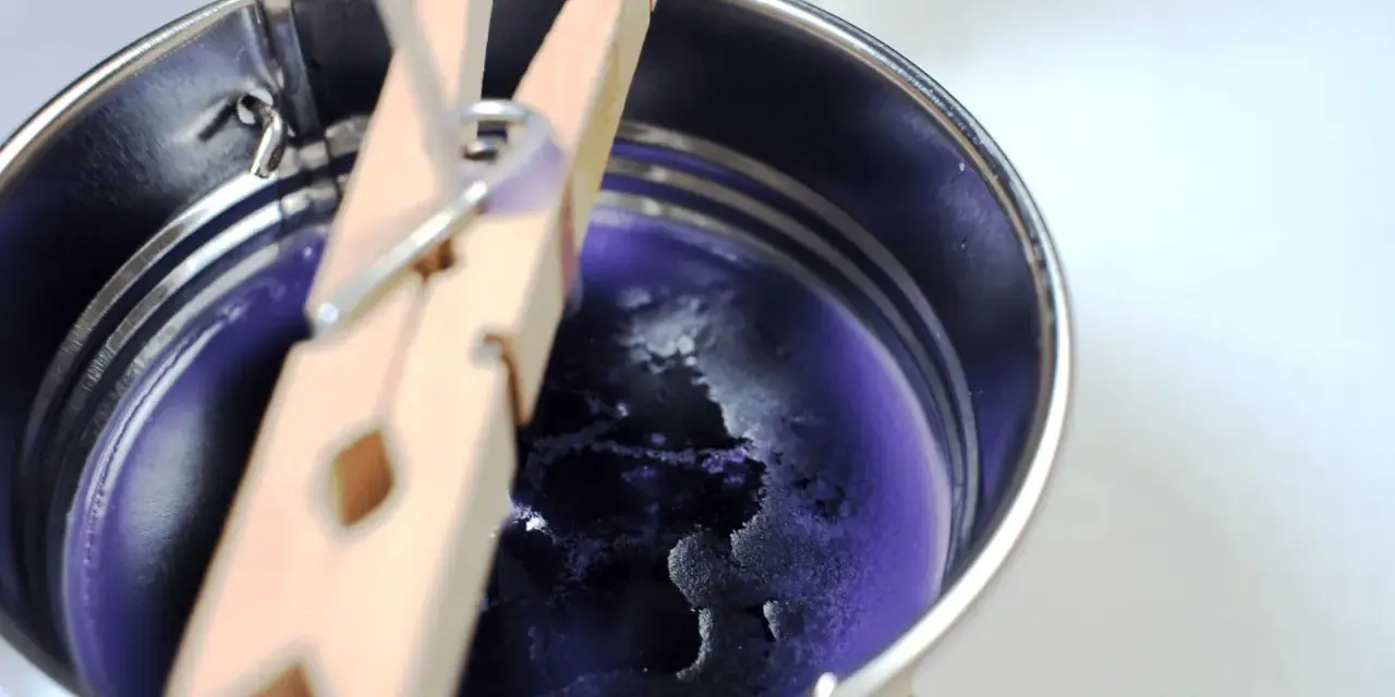 Proceso para como hacer velas de soja: cera morada enfriándose en un recipiente metálico, sujeta por pinzas de madera.