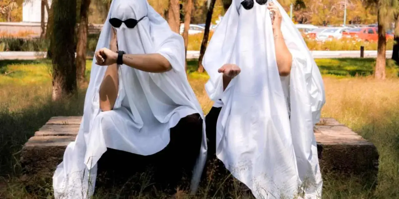 Dos figuras cubiertas con sábanas blancas y gafas de sol, sentadas en un banco de piedra. Una mira su reloj, la otra extiende la mano. ¡Ideas de terror para un disfraz!