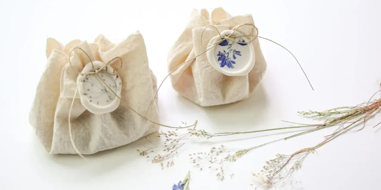 Dos pequeños paquetes de tela rústica, atados con hilo y sellados con cera, decorados con flores secas. Son detalles de boda originales hechos a mano, perfectos para un toque natural.