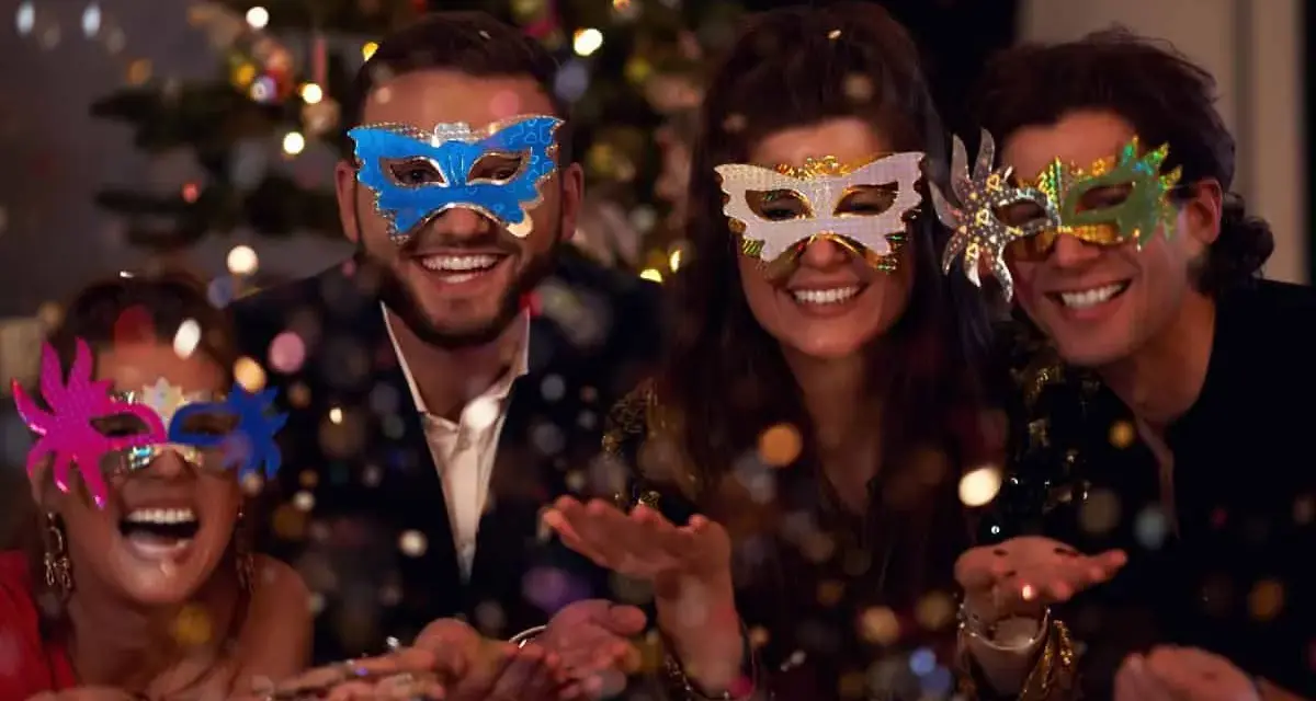 Cuatro amigos disfrutan de una temática fiesta de mascaras, riendo y lanzando confeti frente a un árbol de Navidad iluminado.
