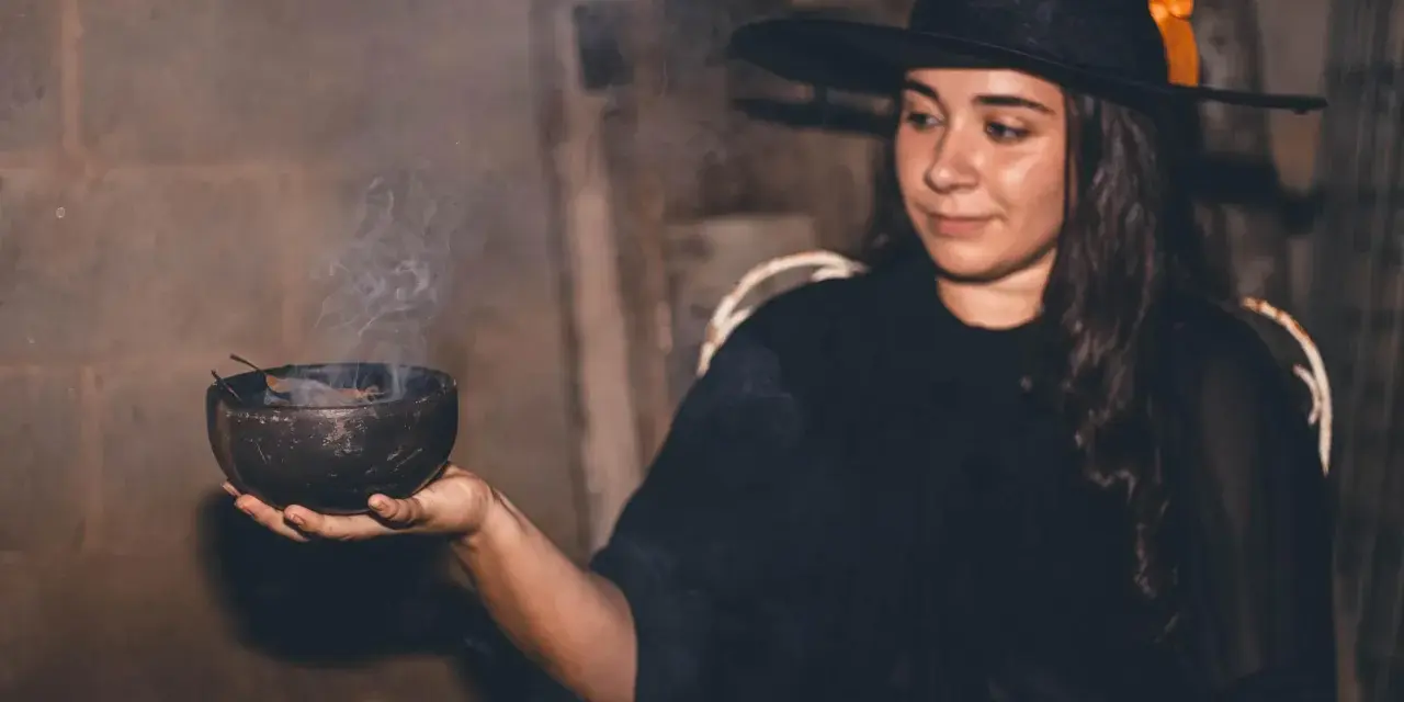 Mujer con disfraz bruja casero adulto, con sombrero negro y naranja, sosteniendo un cuenco humeante.
