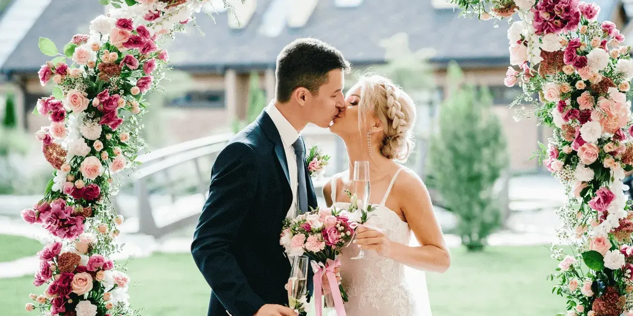 Novios besándose bajo un arco floral rosa y blanco, celebrando su boda barata con copas de champán.