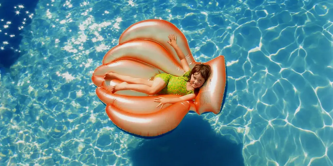 Niña sonriente en un flotador naranja con forma de concha, disfrutando de la pool party y sus juegos bajo el sol.
