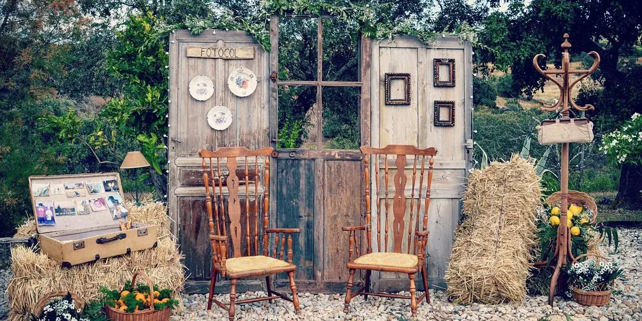 Un rústico photocall boda casero con puertas antiguas, sillas de madera, balas de heno y detalles florales.