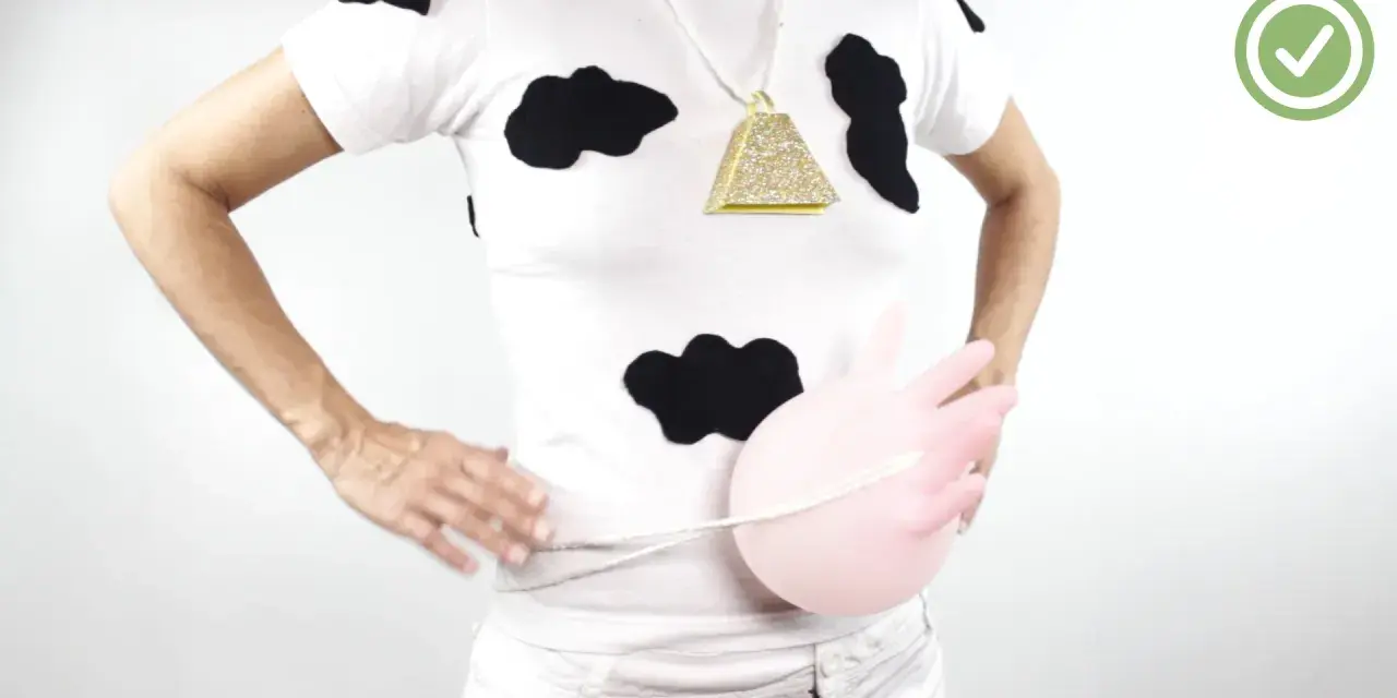 Persona con un disfraz vaca casero: camiseta blanca con manchas negras, collar de campana dorada y un globo rosa simulando la ubre.
