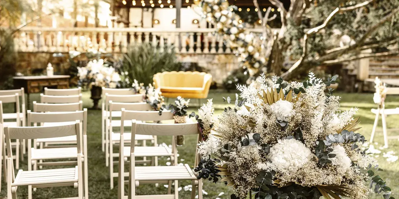 Sillas blancas alineadas en un jardín, un sofá amarillo y un gran arreglo floral. Ideas para como hacer una boda económica y hermosa.