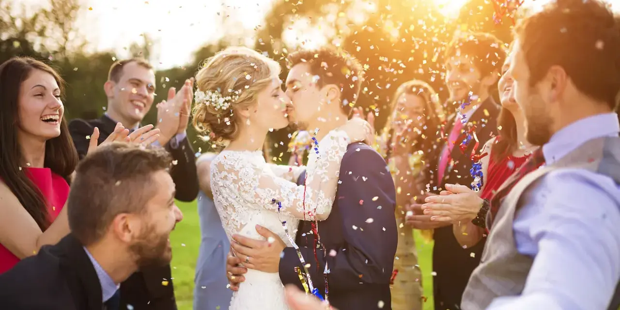 Novios besándose entre confeti mientras amigos aplauden y ríen. ¡Un momento perfecto para felicitar a unos novios!