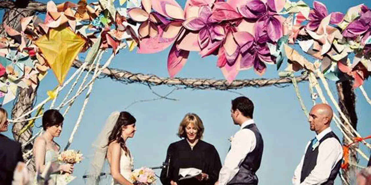 Boda al aire libre con un arco decorado con manualidades de papel coloridas. La pareja se da el sí ante un oficiante.