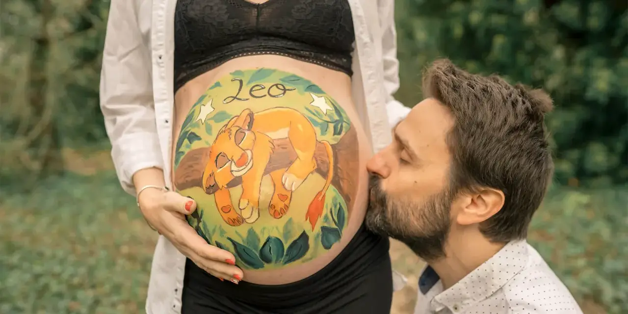 Barriga de embarazada pintada con un león y el nombre "Leo". El padre besa la barriga. Unas plantillas para pintar barriga de embarazada niño, como esta, son un recuerdo precioso.