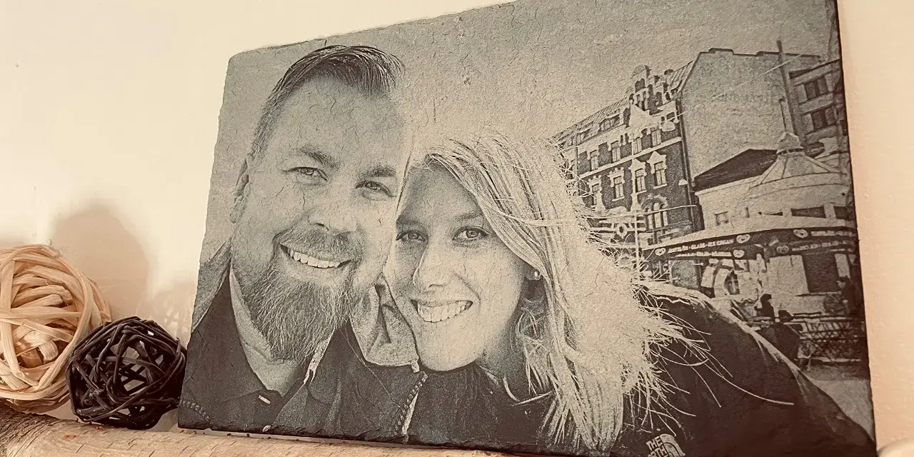 Foto piedra con retrato de pareja sonriente, con edificios y un puesto de helados al fondo.