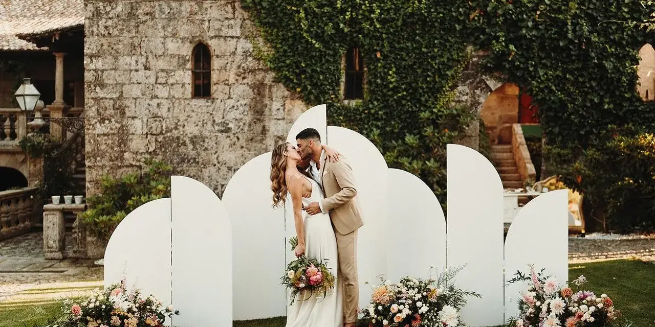 Pareja besándose frente a un photocall bodas de oro casero, decorado con flores y un fondo arquitectónico.