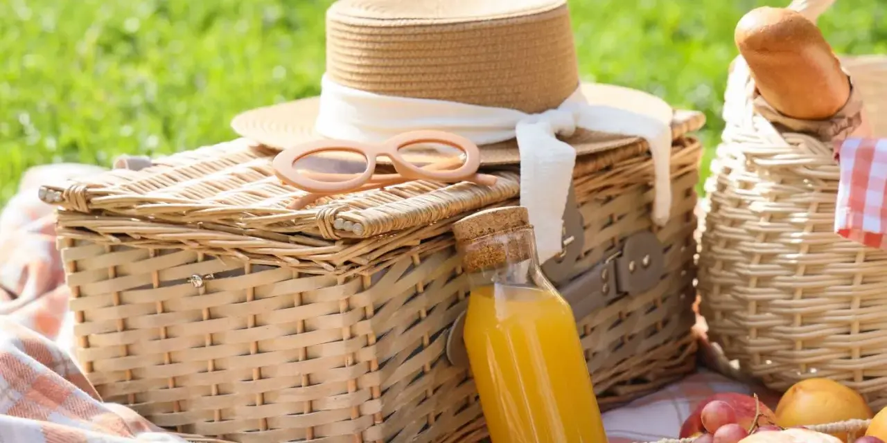 Cajas picnic listas para disfrutar: sombrero, gafas de sol, jugo y frutas sobre mantas a cuadros en un día soleado.