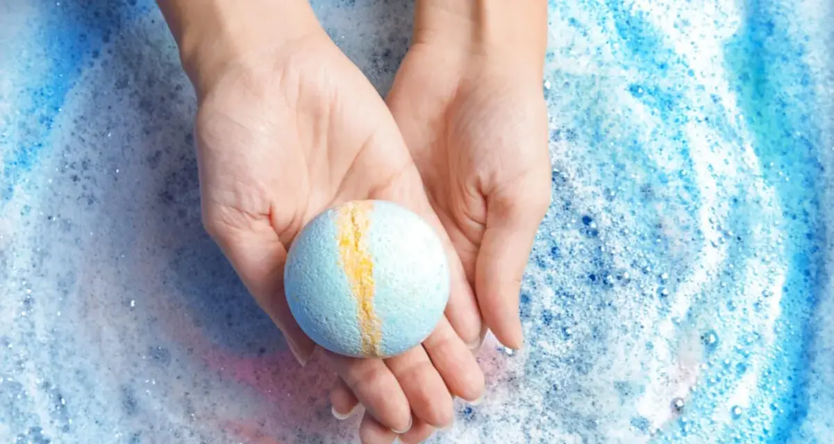 Manos sostienen una bomba de baño azul y amarilla sobre espuma burbujeante. ¡Perfecto para hacer bombas de baño caseras!