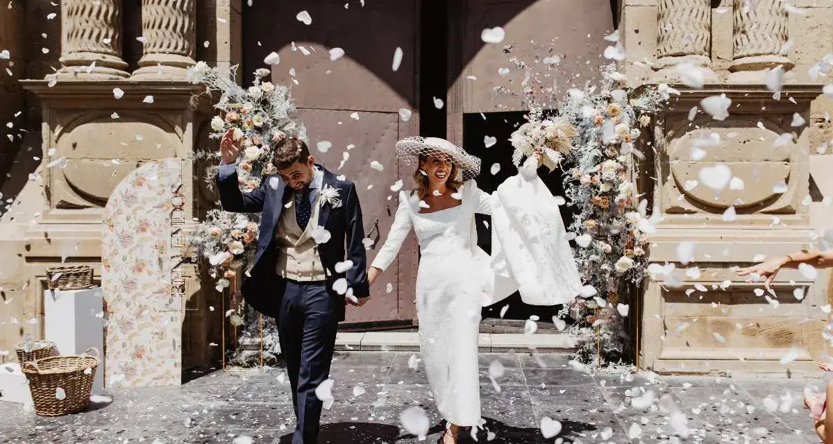 Novios felices salen de la iglesia, cubiertos de pétalos blancos. Es un momento perfecto para tirar confeti en una boda.