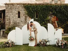 Pareja besándose frente a un photocall bodas de oro casero, decorado con flores y un fondo arquitectónico.