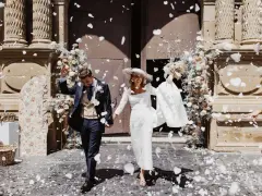 Novios felices salen de la iglesia, cubiertos de pétalos blancos. Es un momento perfecto para tirar confeti en una boda.