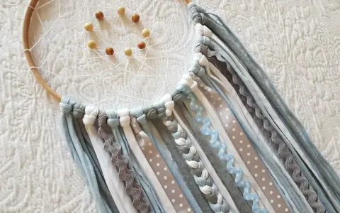 Un atrapasueños casero con aros de madera, red blanca y cuentas. Cintas de tela y encaje cuelgan en tonos grises, blancos y azules.