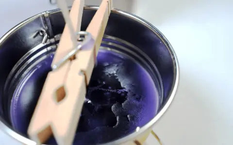 Proceso para como hacer velas de soja: cera morada enfriándose en un recipiente metálico, sujeta por pinzas de madera.