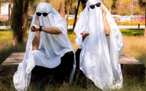Dos figuras cubiertas con sábanas blancas y gafas de sol, sentadas en un banco de piedra. Una mira su reloj, la otra extiende la mano. ¡Ideas de terror para un disfraz!