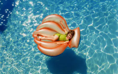 Niña sonriente en un flotador naranja con forma de concha, disfrutando de la pool party y sus juegos bajo el sol.