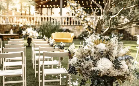 Sillas blancas alineadas en un jardín, un sofá amarillo y un gran arreglo floral. Ideas para como hacer una boda económica y hermosa.