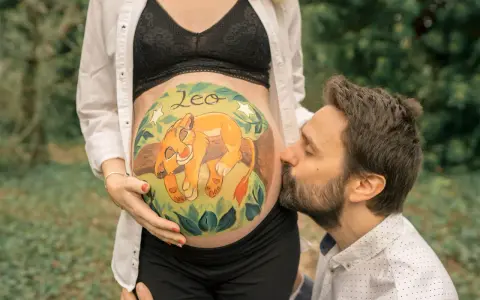 Barriga de embarazada pintada con un león y el nombre "Leo". El padre besa la barriga. Unas plantillas para pintar barriga de embarazada niño, como esta, son un recuerdo precioso.