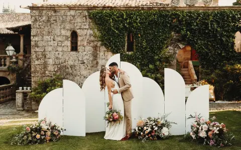 Pareja besándose frente a un photocall bodas de oro casero, decorado con flores y un fondo arquitectónico.