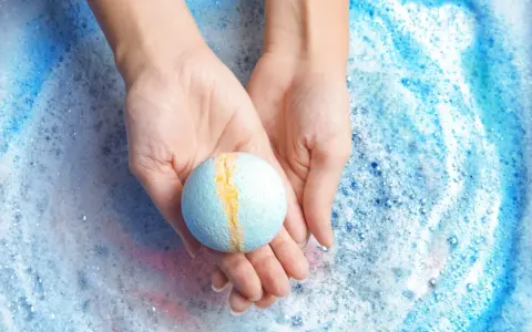 Manos sostienen una bomba de baño azul y amarilla sobre espuma burbujeante. ¡Perfecto para hacer bombas de baño caseras!