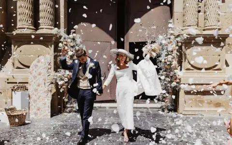 Novios felices salen de la iglesia, cubiertos de pétalos blancos. Es un momento perfecto para tirar confeti en una boda.
