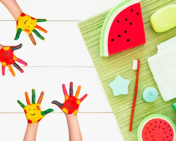 Manos pintadas de colores con caritas sonrientes, listas para hacer jabones para niños. Hay jabones con forma de sandía y otros accesorios de baño.