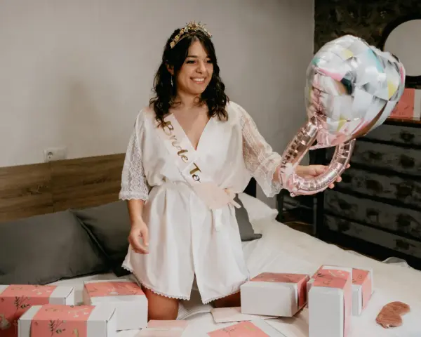 Novia sonriente con corona y banda "Bride To Be", sosteniendo un globo de anillo. Alrededor, cajas de regalo y un pack de accesorios para despedida de soltera.