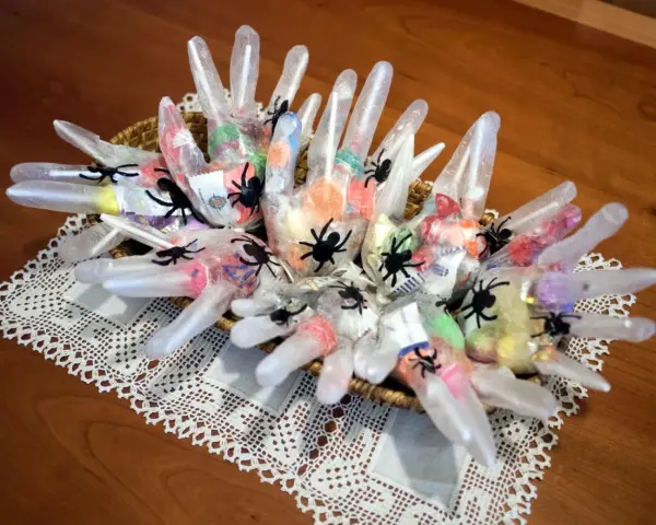 Manualidades chuches Halloween: guantes transparentes rellenos de caramelos y decorados con arañas negras, presentados en una cesta sobre un mantel de encaje.