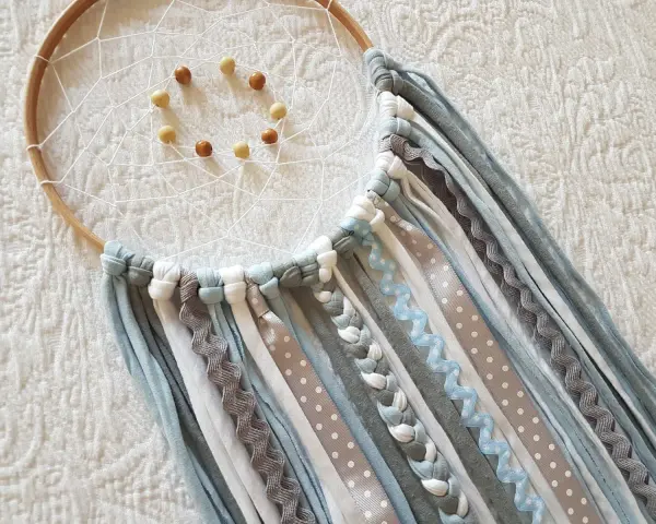 Un atrapasueños casero con aros de madera, red blanca y cuentas. Cintas de tela y encaje cuelgan en tonos grises, blancos y azules.