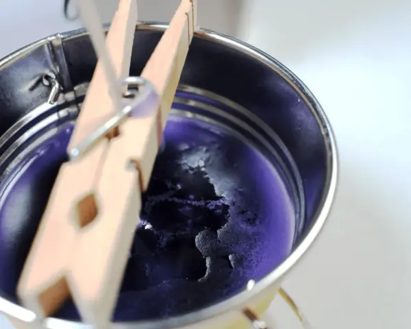 Proceso para como hacer velas de soja: cera morada enfriándose en un recipiente metálico, sujeta por pinzas de madera.