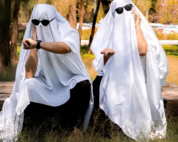 Dos figuras cubiertas con sábanas blancas y gafas de sol, sentadas en un banco de piedra. Una mira su reloj, la otra extiende la mano. ¡Ideas de terror para un disfraz!
