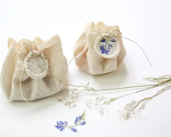 Dos pequeños paquetes de tela rústica, atados con hilo y sellados con cera, decorados con flores secas. Son detalles de boda originales hechos a mano, perfectos para un toque natural.