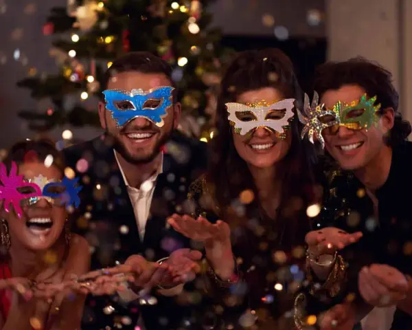Cuatro amigos disfrutan de una temática fiesta de mascaras, riendo y lanzando confeti frente a un árbol de Navidad iluminado.