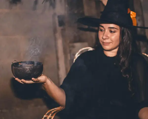 Mujer con disfraz bruja casero adulto, con sombrero negro y naranja, sosteniendo un cuenco humeante.