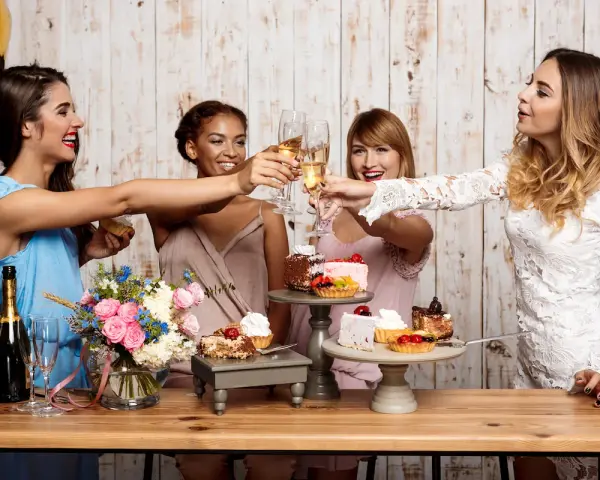 Cuatro amigas brindan con copas de champán en unas fiestas en casa, rodeadas de pasteles y flores.